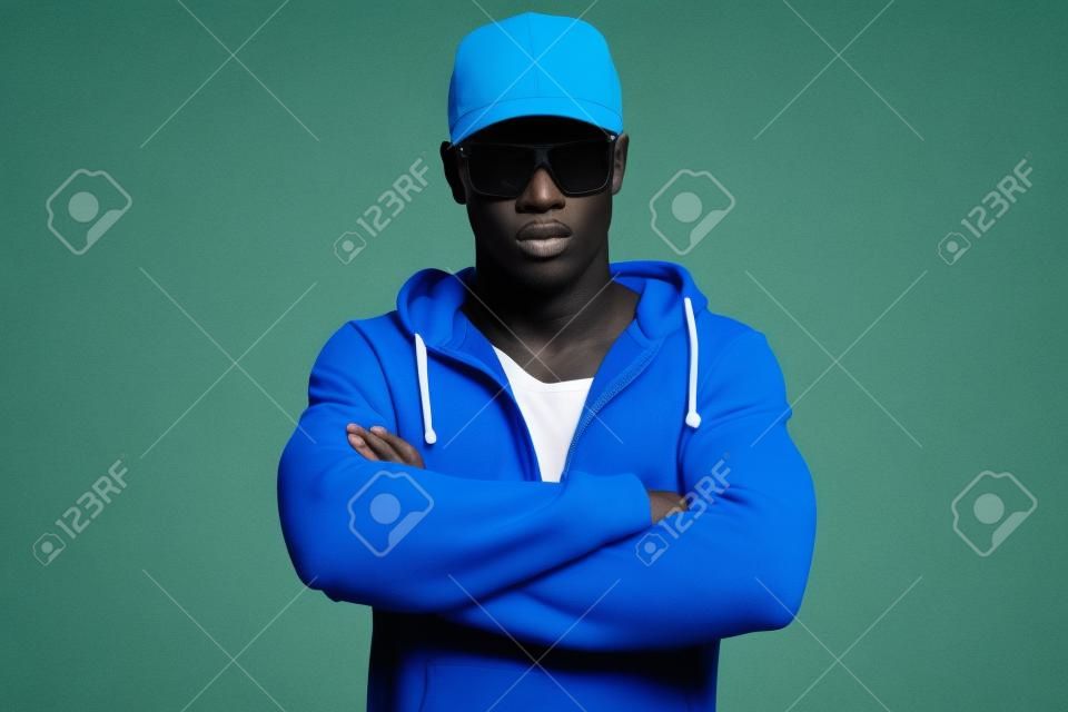 Athletisch Läufer mit Sonnenbrille mit blauen Sportmode. Schwarzer Mann. Blaue Mütze und Pullover. Intensive Farben. Studioaufnahme vor gelbem Hintergrund.