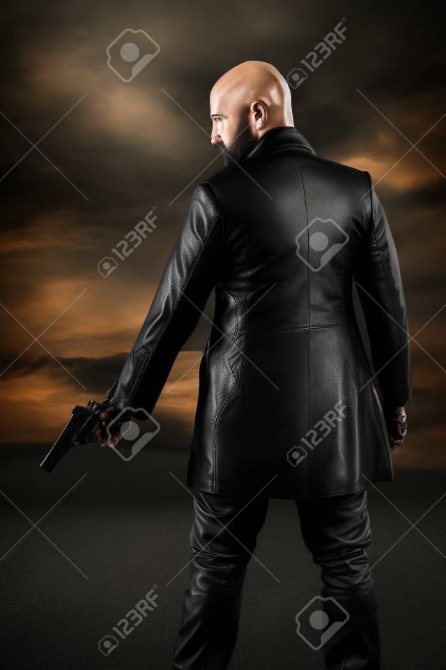 Homem de gângster careca perigoso com arma de retenção de barba. Vestindo jaqueta de couro preto. Céu nublado escuro.