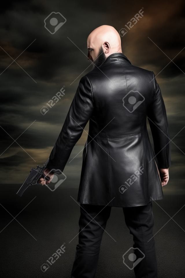 Hombre peligroso gángster calvo con barba sosteniendo la pistola. El uso de la chaqueta de cuero negro. Cielo nublado oscuro.