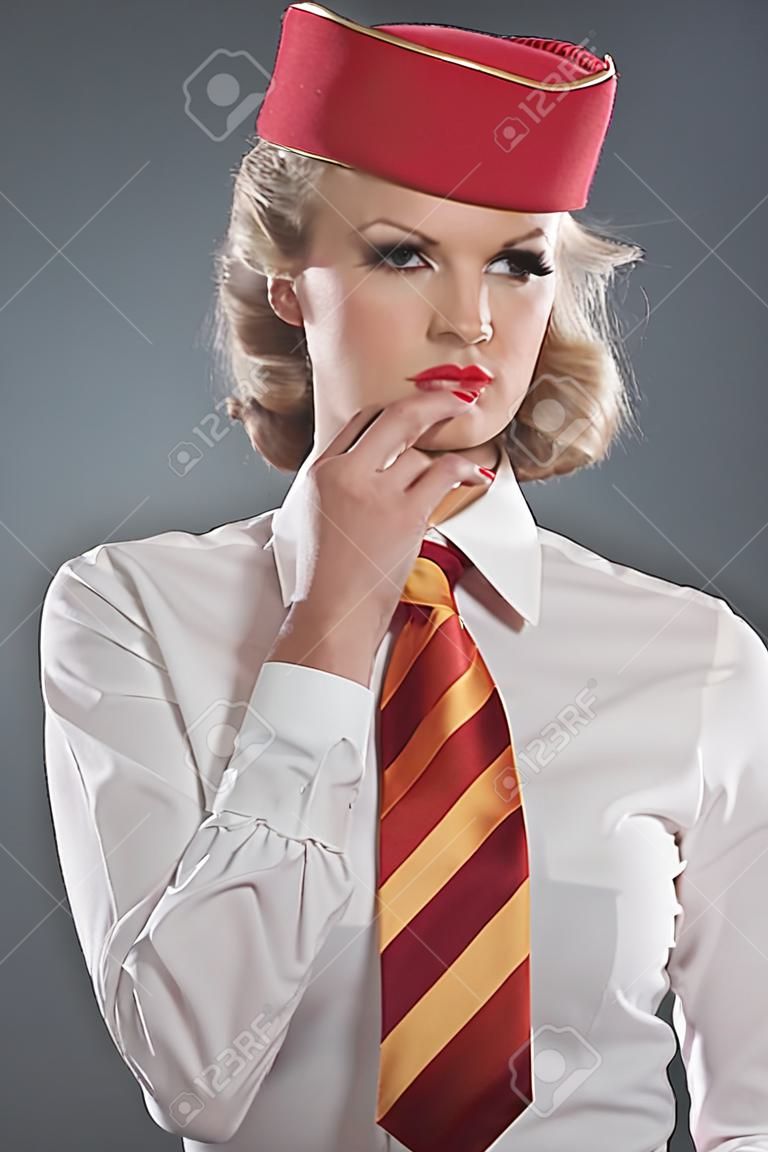 Serious retro blonde stewardess wearing red cap with striped tie and white shirt. Studio shot against grey.