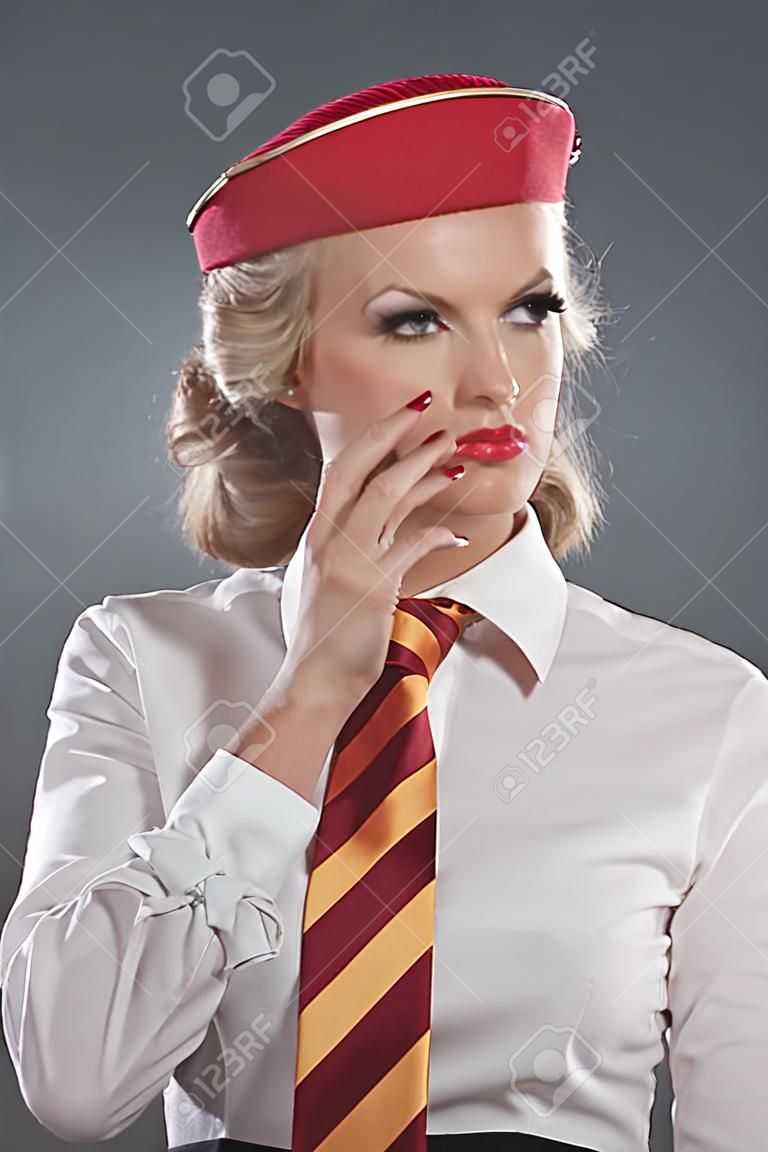 Serious retro blonde stewardess wearing red cap with striped tie and white shirt. Studio shot against grey.