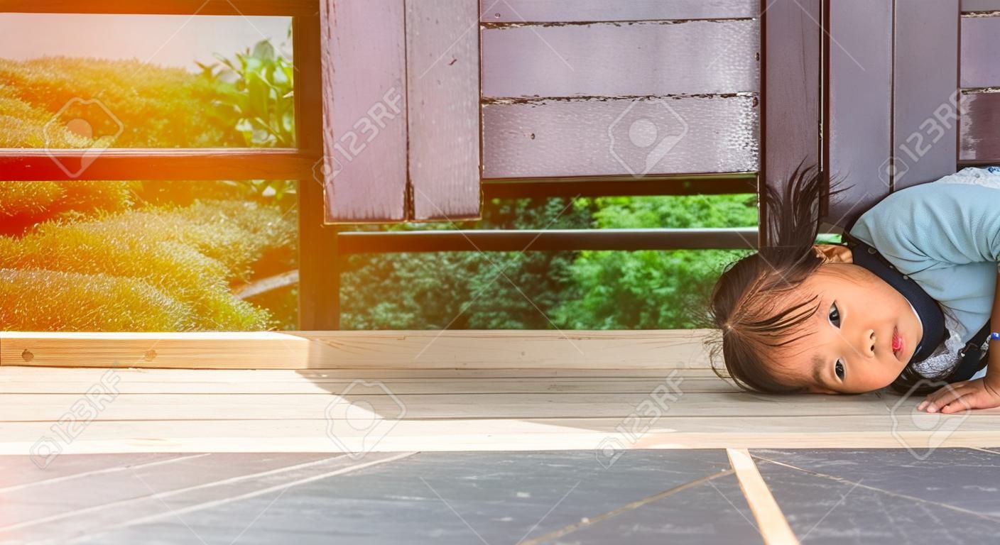 Happy Asian little child girl plays peekaboo under the wooden door in the garden.