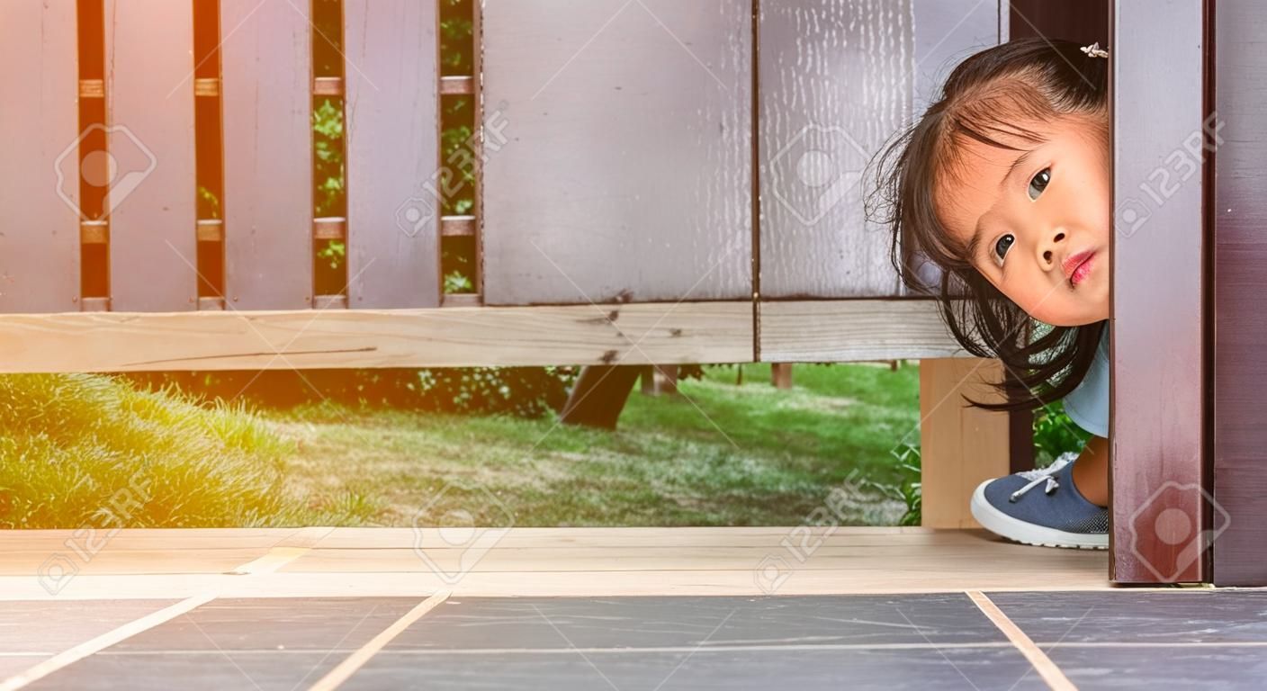 Una bambina asiatica felice gioca a cucù sotto la porta di legno in giardino.