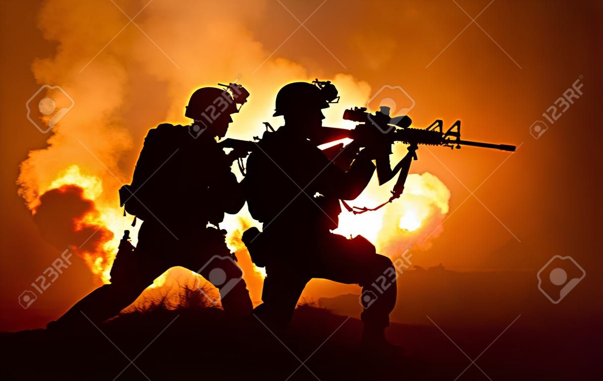 Silhouettes of two army soldiers, U.S. marines team in action, surrounded fire and smoke, shooting with assault rifle and machine gun, attacking enemy with suppressive gunfire during offensive mission