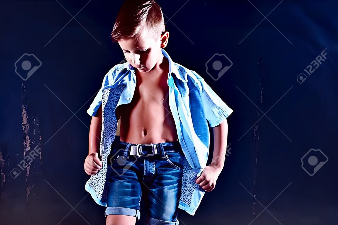 Menino de shorts posando para a câmera e mostrando seu abdômen enquanto está em pé no fundo da parede escura.