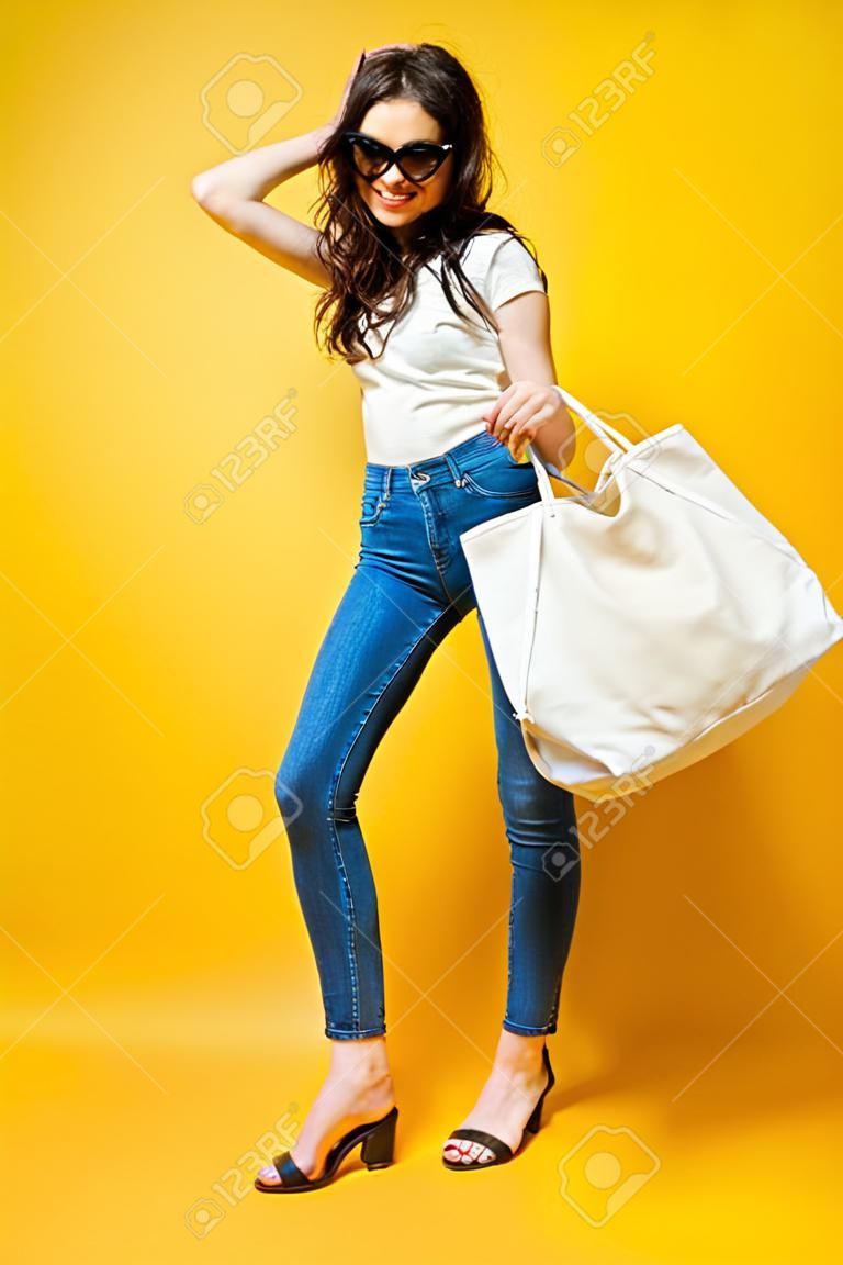 Piękna młoda kobieta w okularach przeciwsłonecznych, białej koszuli, niebieskich dżinsach pozowanie z torbą na żółtym tle