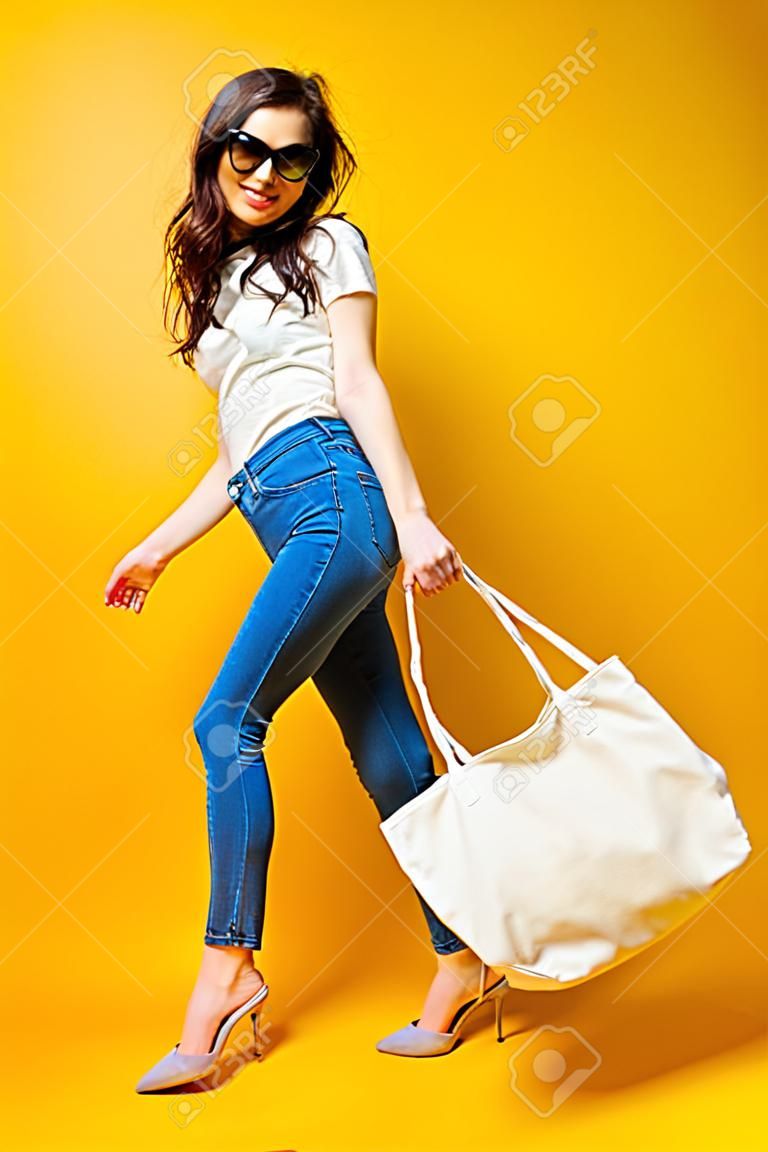 Piękna młoda kobieta w okularach przeciwsłonecznych, białej koszuli, niebieskich dżinsach pozowanie z torbą na żółtym tle