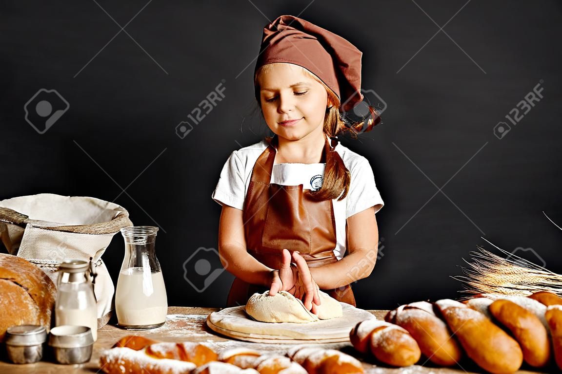 Charmantes kleines Mädchen in Schürze und Hut, das am Tisch steht, Brotteig knetet und Spaß beim Kochen hat