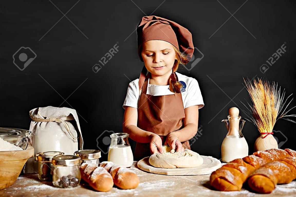 Charmantes kleines Mädchen in Schürze und Hut, das am Tisch steht, Brotteig knetet und Spaß beim Kochen hat