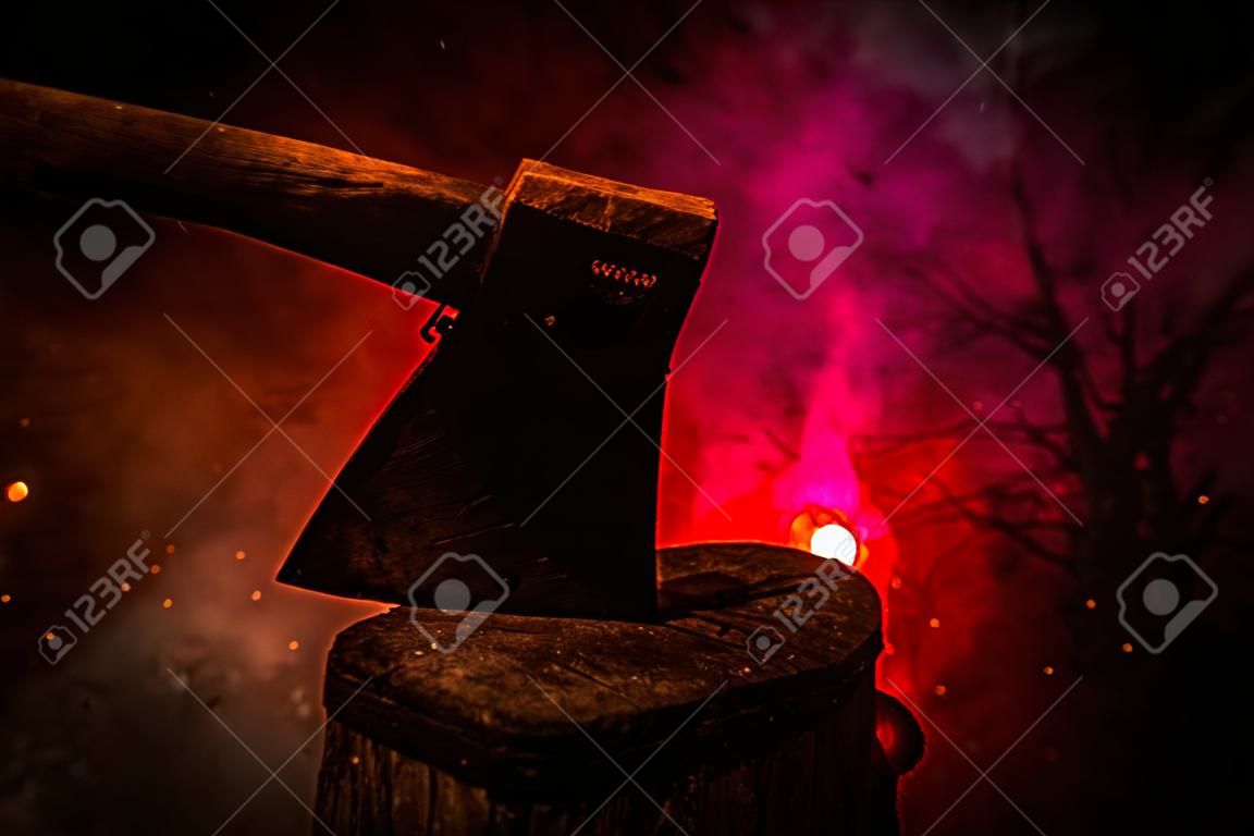 Old ax attached to the tree trunk on horror red foggy background. Scary Halloween theme with maniac killer weapon.