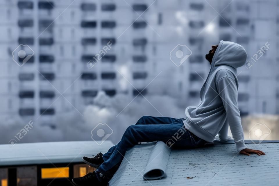 Mysterie man in wit masker dragen hoody zittend op het dak van verlaten gebouw en kijken omhoog in de lucht voelen eenzaam, zelfvernietiging suïcidale verslaving of grote depressieve wanorde concepten