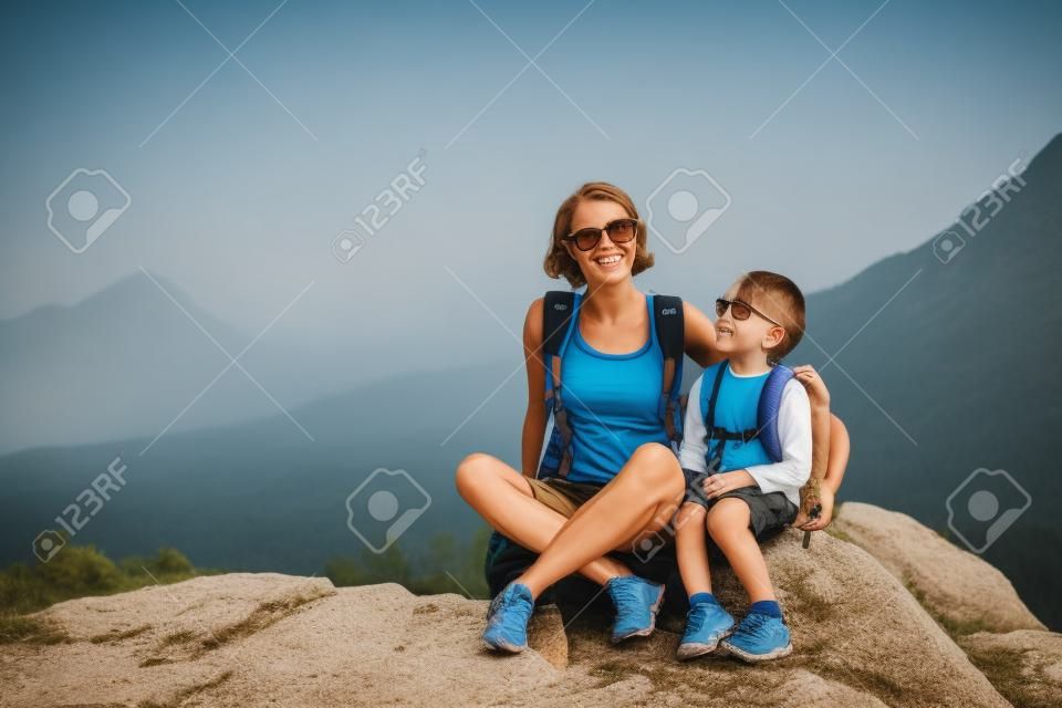 少年と彼の母親は山の頂上に座っています。女性は子供と一緒に旅行します。赤ちゃんはお母さんを抱きしめる。バックパック付きの旅。ハイキングや子供たちと一緒に登ります。息子と一緒に女性の肖像画。
