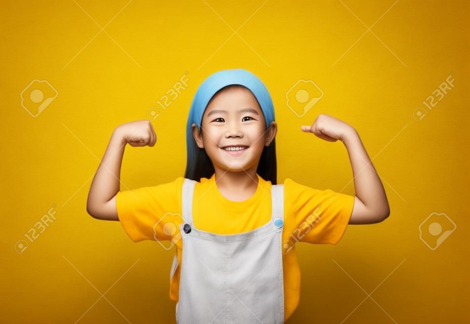 Menina asiática sorridente que mostra seu músculo com a câmera olhando isolada no fundo amarelo.