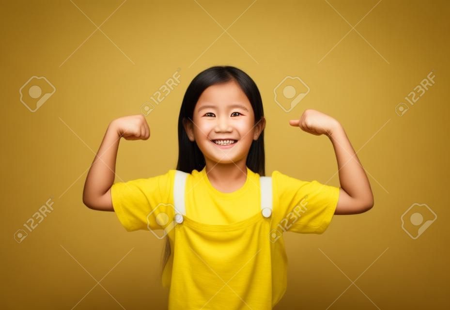 Menina asiática sorridente que mostra seu músculo com a câmera olhando isolada no fundo amarelo.