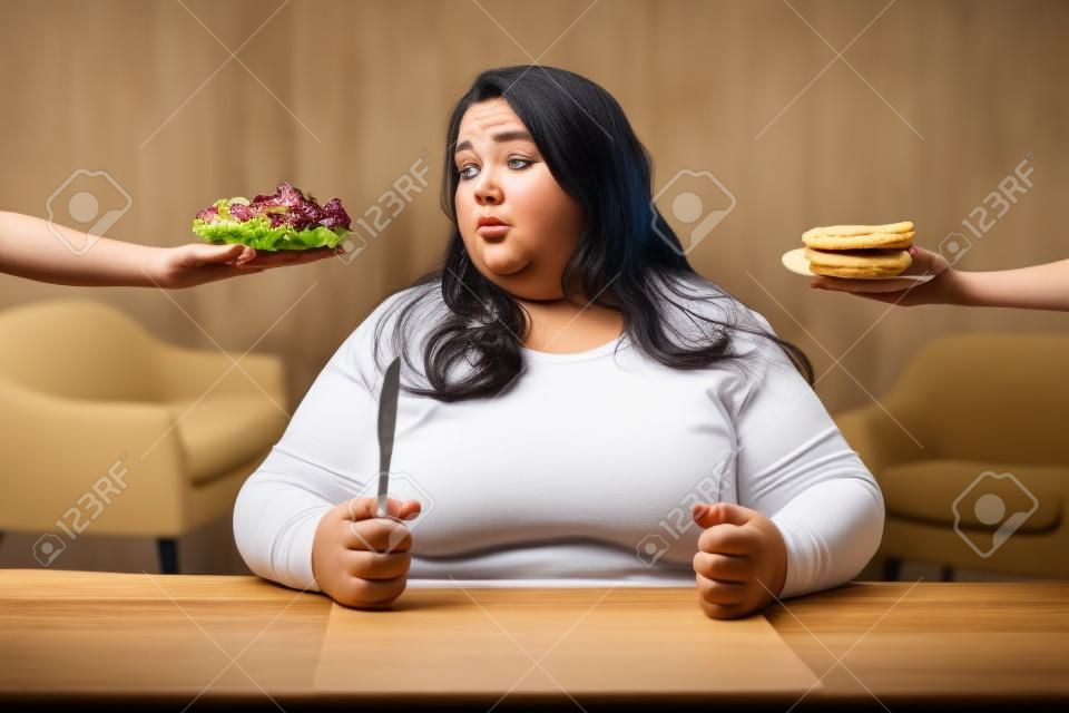 Fett sein. Elende pralle Frau, die einen Salat betrachtet und Kekse will