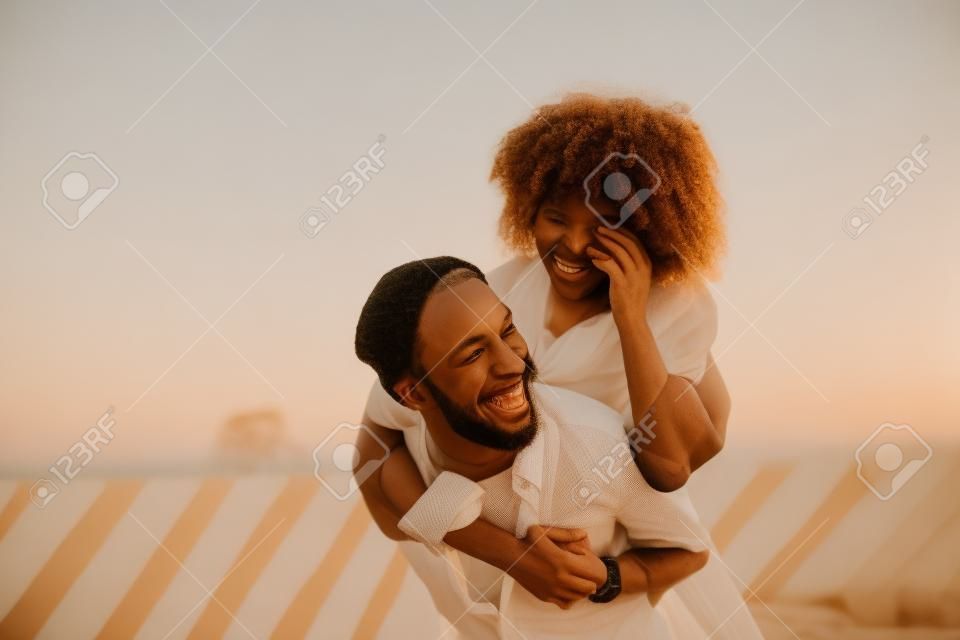 On the back. Cheerful young man smiling while carrying his beloved young woman on his back