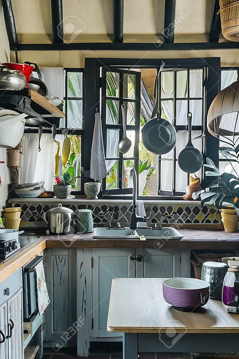 Old crampy rustic kitchen with small window in a tropical house with straw roof. Old furniture, shelves covering every millimeter of a wall space with cooking ware, tableware and food on them.