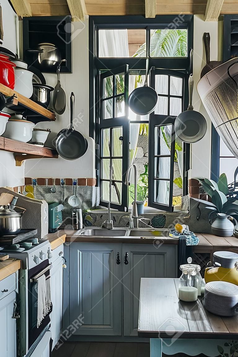 Old crampy rustic kitchen with small window in a tropical house with straw roof. Old furniture, shelves covering every millimeter of a wall space with cooking ware, tableware and food on them.